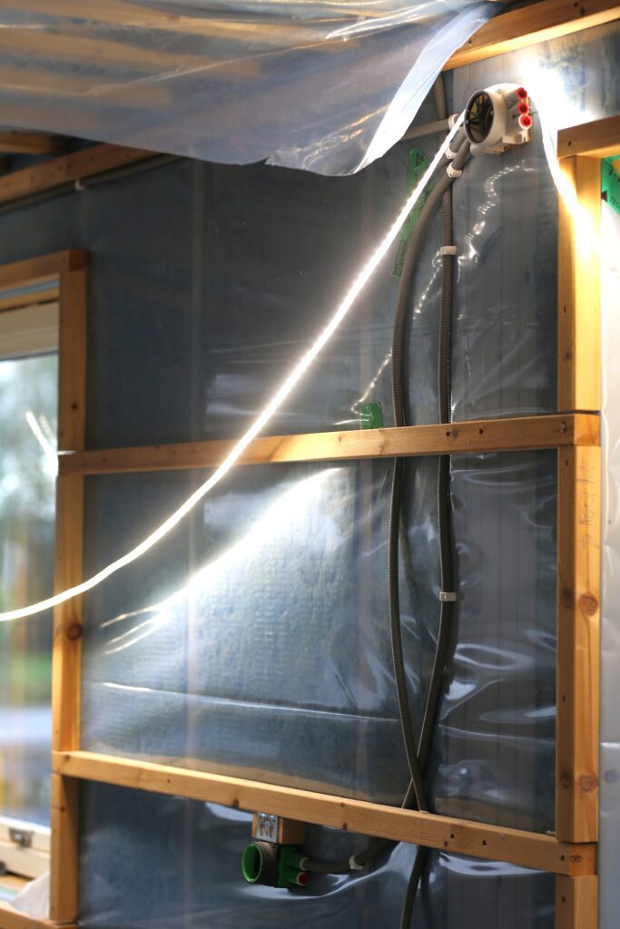 Att bygga hus - Öppet installationsskikt med byggbelysning, synliga rör och kablar vid fönster och altandörr under husbygge.